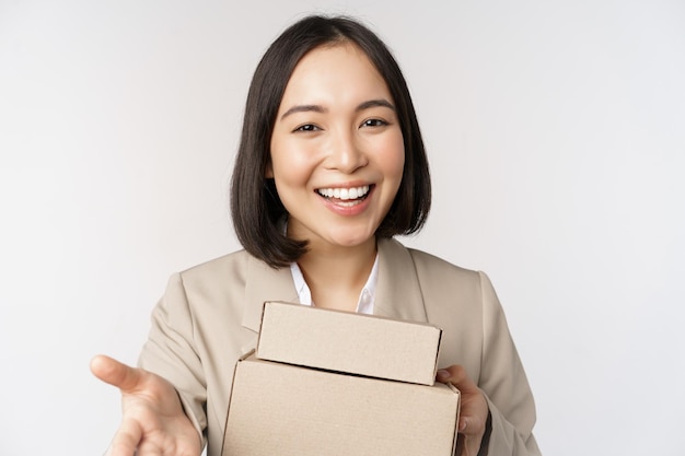 Free photo portrait of business woman asian saleswoman pointing at you giving boxes with orders standing in suit over white background