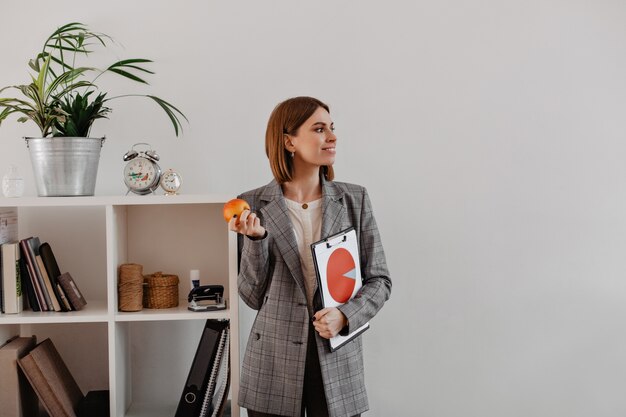 Portrait of business woman with pie chart in her hands looking to side. Smiling woman is going to eat apple for lunch in office.