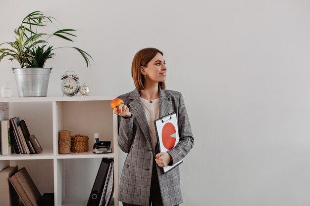Портрет деловой женщины с круговой диаграммой в руках, глядя в сторону. Улыбающаяся женщина собирается съесть яблоко на обед в офисе.
