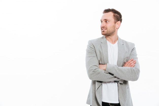 Portrait of businesslike man in jacket posing with broad smile keeping arms folded and looking aside, isolated over white wall copy space