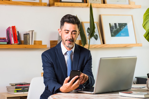 Ritratto dell'uomo d'affari con il computer portatile sulla sua tavola facendo uso del telefono cellulare