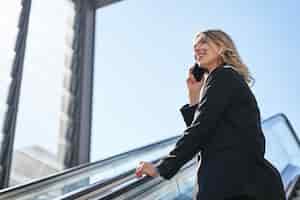 Free photo portrait of businesswoman in black suit going up on escalator talking on mobile phone saleswoman wal