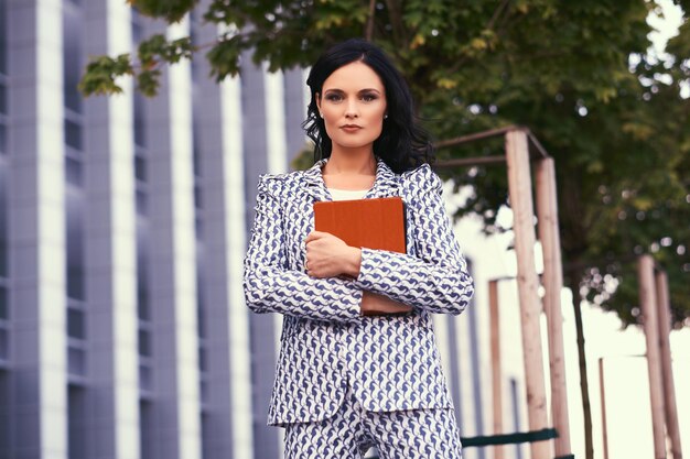 Portrait of businesswoman dressed in a stylish suit, holds a tablet computer, standing in a downtown against a background of a skyscraper.