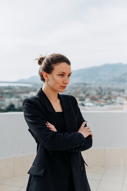 Portrait of businesswoman at rooftop standing with hands crossed and thinking in black suit during daytime.