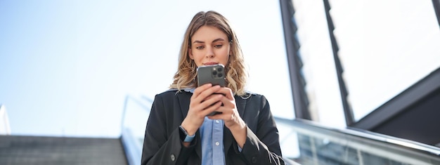 Free photo portrait of businesswoman in suit standing on escalator walking in city using mobile phone corporate