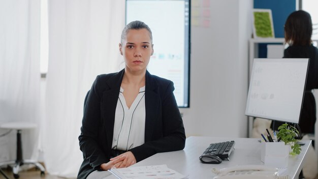 Portrait of businesswoman with office job sitting at desk with computer monitor and data charts on screen. Corporate employee looking at camera and preparing to work on business project