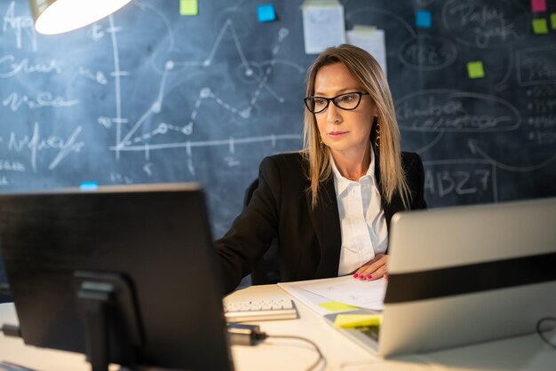 Портрет деловой женщины в рабочем процессе. Серьезная женщина, работающая с двумя компьютерами по вечерам, анализирует финансовые данные, рассчитывает прибыль для инвестиций. Концепция роста бизнеса и инвестиционной деятельности