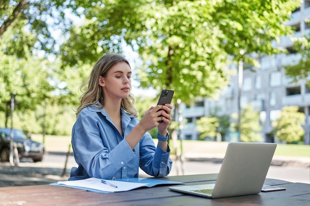 Portrait of businesswoman working outside in park sitting with laptop work documents and using mobil