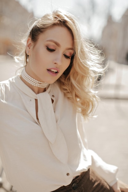 Portrait of calm blonde curly woman in pearl necklace white blouse and velvet brown pants poses outside