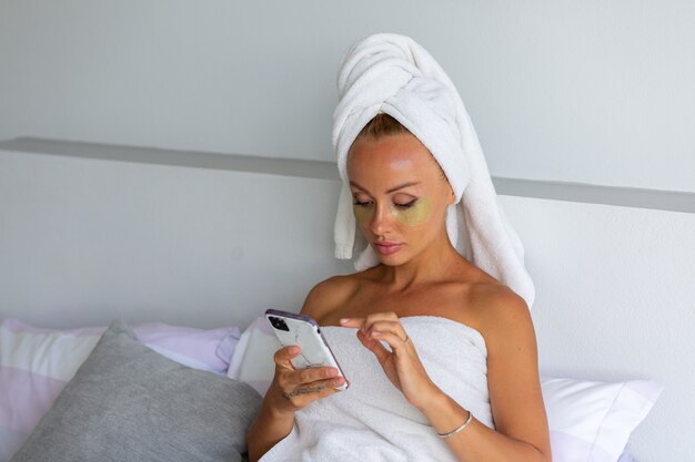 Portrait of calm caucasian pretty woman with a towel on head and eye mask patches on face Face skin care concept Female relax on bed at home take selfie watch phone