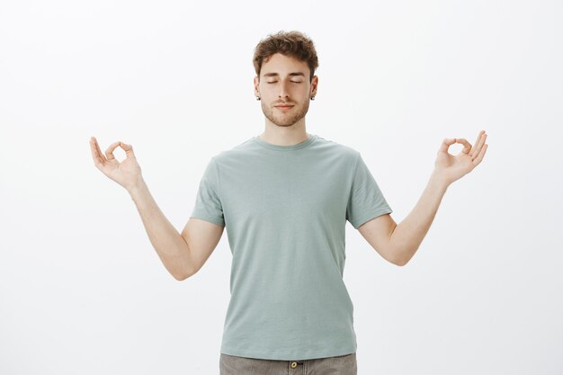 Portrait of calm good-looking Caucasian man in t-shirt, smiling and feeling relaxed, standing with spread hands in zen gesture and closed eyes