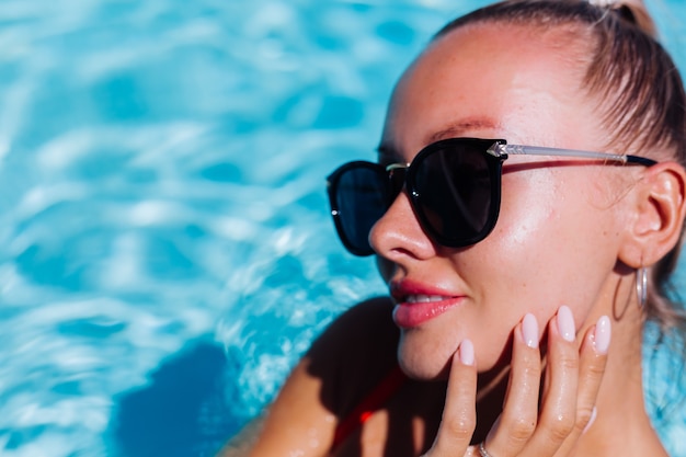 Portrait of calm happy woman in sunglasses with tanned skin in blue swimming pool at sunny day