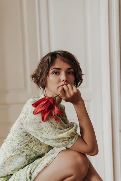 Портрет спокойной молодой брюнетки с короткими волосами в цветочном платье, опираясь на колени, смотрит вперед и держит красный цветок