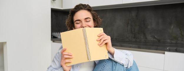 Free photo portrait of candid smiling woman cover her face with notebook laughing carefree doing her homework