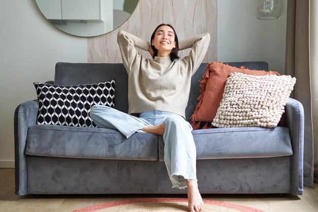 Portrait of carefree asian woman enjoying dayoff sitting on sofa and smiling pleased relaxing in hou