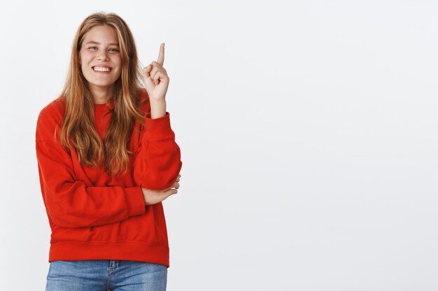 Portrait of carefree charismatic woman rembemer cool joke raising index finger while laughing and smiling adding word with eureka gesture giving suggestion or idea posing in red sweater over grey wall