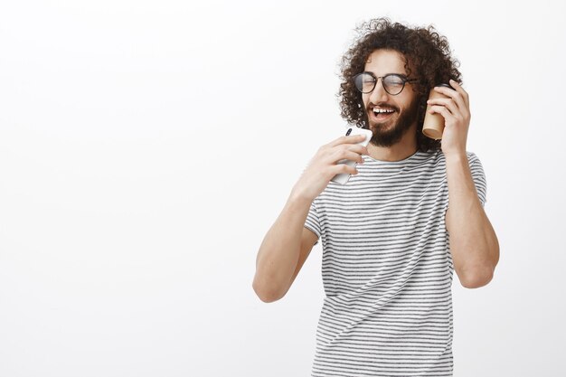 Portrait of carefree gunny handsome man with beard in black glasses, holding smartphone like microphone and singing along with cup of coffee near ear