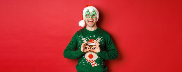 Portrait of carefree handsome man in santa hat and party glasses, making fun of his christmas sweater, looking happy over red background