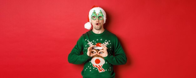 Portrait of carefree handsome man in santa hat and party glasses making fun of his christmas sweater...