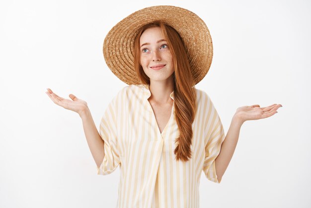 Portrait of carefree optimistic charming kind female with ginger hair and freckles in cute straw hat and yellow blouse making shoulder shrugg with hands spread and innocent look