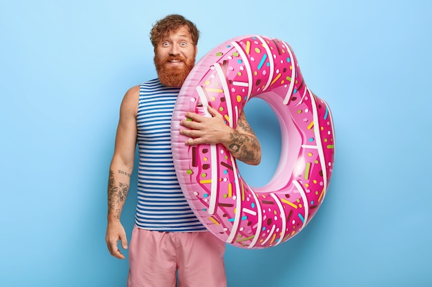 Free photo portrait of carefree smiling red haired man posing with donut pool floaty