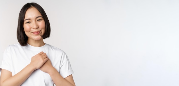 Portrait of carig young asian woman holding hands on heart gazing with care and love touched by something feel flattered standing over white background