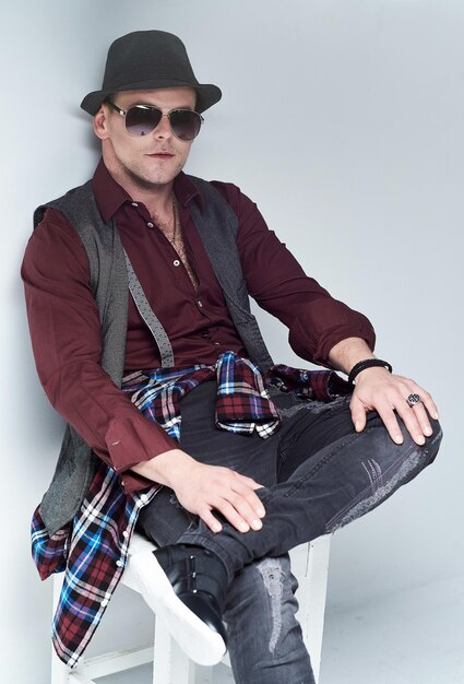 Portrait of casual male dressed in a shirt, waistcoat and sunglasses sits on a chair.