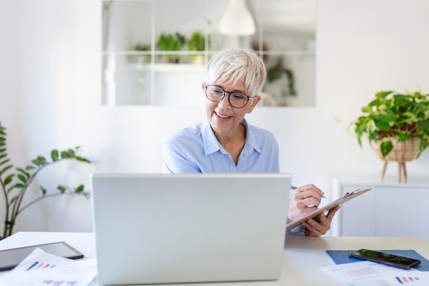 사무실에 앉아 일하는 동안 노트북을 사용하는 캐주얼 여성의 초상화 노트북 앞에 앉아 집에서 소규모 비즈니스를 관리하는 매력적인 중년 여성