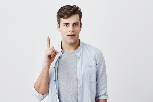 Portrait of caucasian bug-eyed male with dark hair being shocked by something, dressed in blue shirt over t-shirt, pointing his finger at blank wall with copy space for your advertising content