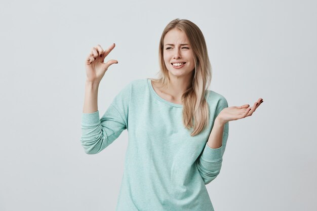 portrait of caucasian female model with blonde hair clenching her teeth with puzzlement, demonstrating size of something small in size gesturing actively. Body language and face expression