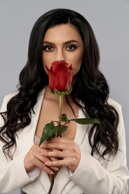 Портрет кавказской женской модели с темными волосами, скрывающими губы за красной розой, которая держится в руках. Очаровательная молодая женщина позирует в студии с серым фоном.