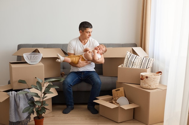 Portrait of caucasian man wearing white t-shirt and jeans sitting on sofa with his baby daughter, moving to a new flat or house, carrying crying child, trying to calm down.