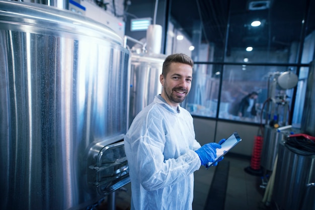 Portrait of caucasian man in white suit standing by the reservoirs in production plant Industrial worker with tablet