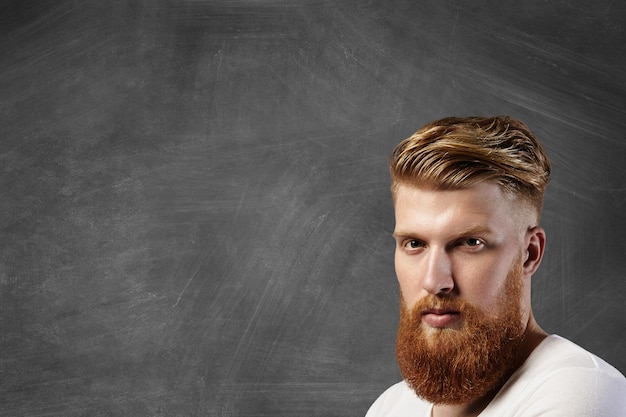 Free photo portrait of caucasian man with stylish haircut and big fuzzy beard wearing white t-shirt, having thoughtful pensive expression while standing against blank chalkboard