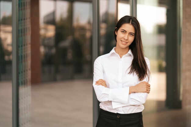 Portrait of a Caucasian pretty business lady smiling and crossing her hands while standing on the glass building