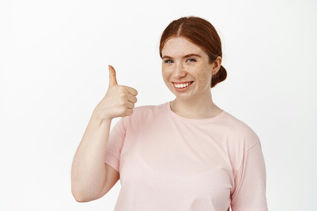 Portrait of caucasian redhead girl shows thumb up and smiles pleased, support your idea, praise and compliment, like something good, positive feedback, standing on white