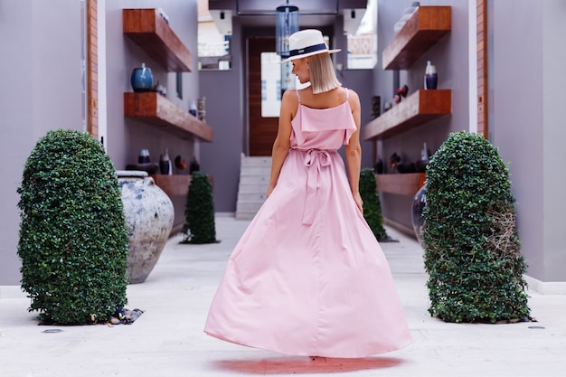 Portrait of caucasian woman in romantic elegant pink long dress on vacation at luxury rich villa hotel with amazing tropical palm trees view Female in classic white hat