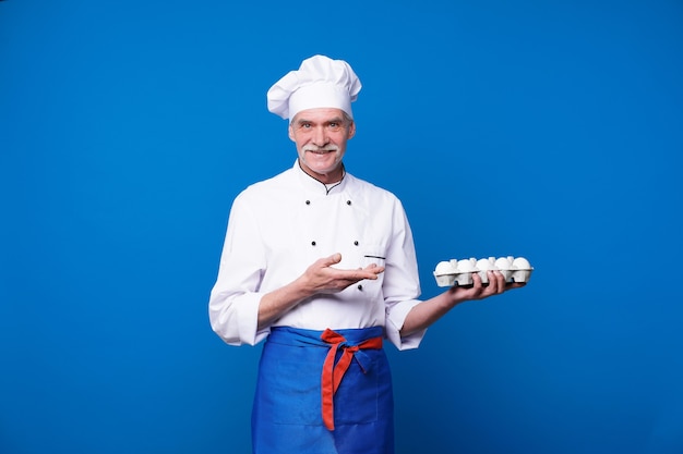 Portrait of charismatic bearded chef holding basket with fresh eggs while posing against blue wall