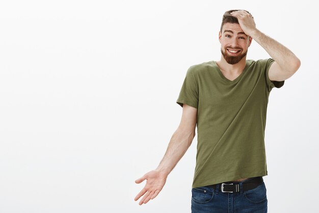 Portrait of charismatic handsome bearded man apologizing for being late, forgetting about time holding hand on head guilty shrugging cute and holding hand sideways, smiling as telling sorry