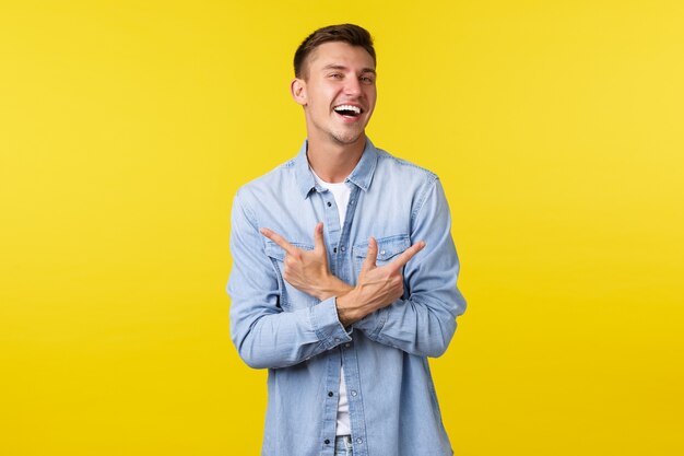 Portrait of charismatic handsome blond man laughing happy and pointing fingers sideways, showing left and right variants or products, standing cheerful over yellow background.