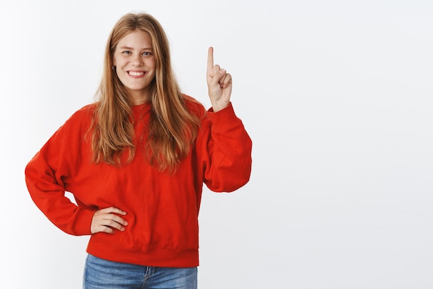 Portrait of charismatic joyful good-looking young caucasian girl, freckles and blue eyes holding hand on waist raising finger and pointing up