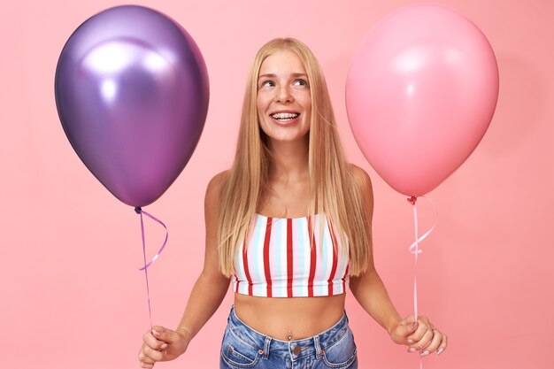 Portrait of charming adorable student girl holding two metallic helium balloons, celebrating birthday, having fun