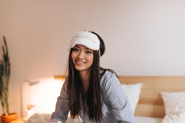 Free photo portrait of charming asian girl in white fluffy sleep mask woman looks into camera and sits on bed