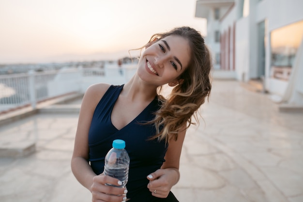 Ritratto affascinante attraente giovane donna in abiti sportivi con bottiglia d'acqua durante l'allenamento in mattina di sole sul lungomare nel paese tropicale. lavoro a domicilio, sorridere, esprimere positività.