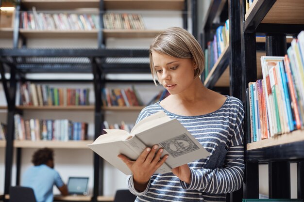 Портрет очаровательной белокурой студентки с короткими волосами в повседневной одежде, стоящей возле полки в библиотеке, читающей книгу, просматривающей информацию об экономических системах.