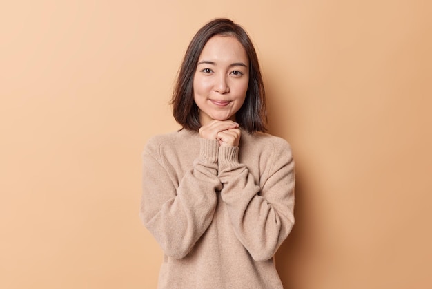 Portrait of charming brunette Asian woman keeps hands under chin smiles gently wears soft warm jumper isolated over beige background. Pretty young female model with clean skin feels satisfied