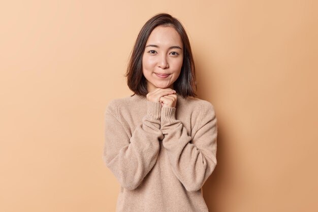 Portrait of charming brunette Asian woman keeps hands under chin smiles gently wears soft warm jumper isolated over beige background. Pretty young female model with clean skin feels satisfied