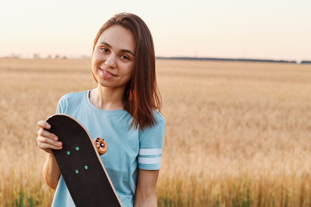 Portrait of charming brunette woman wearing blue t shirt looking directly at camera, holding skateboard in hands, copy space for advertisement, healthy lifestyle.