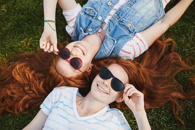 Portrait of charming and carefree redhead female siblings with freckles, lying on grass in park and wearing trendy sunglasses while laughing and smiling, discussing shape of clouds.