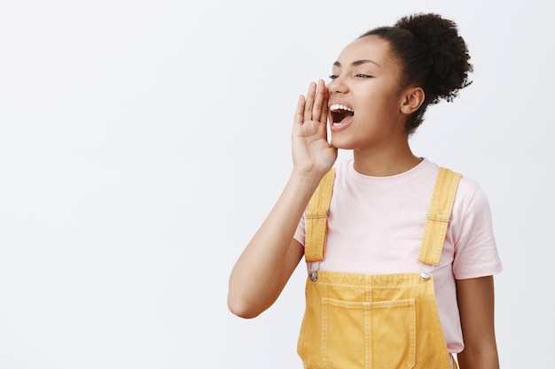 Portrait of charming carefree urban female in yellow trendy overalls, turning left and holding palm near opened mouth, yelling